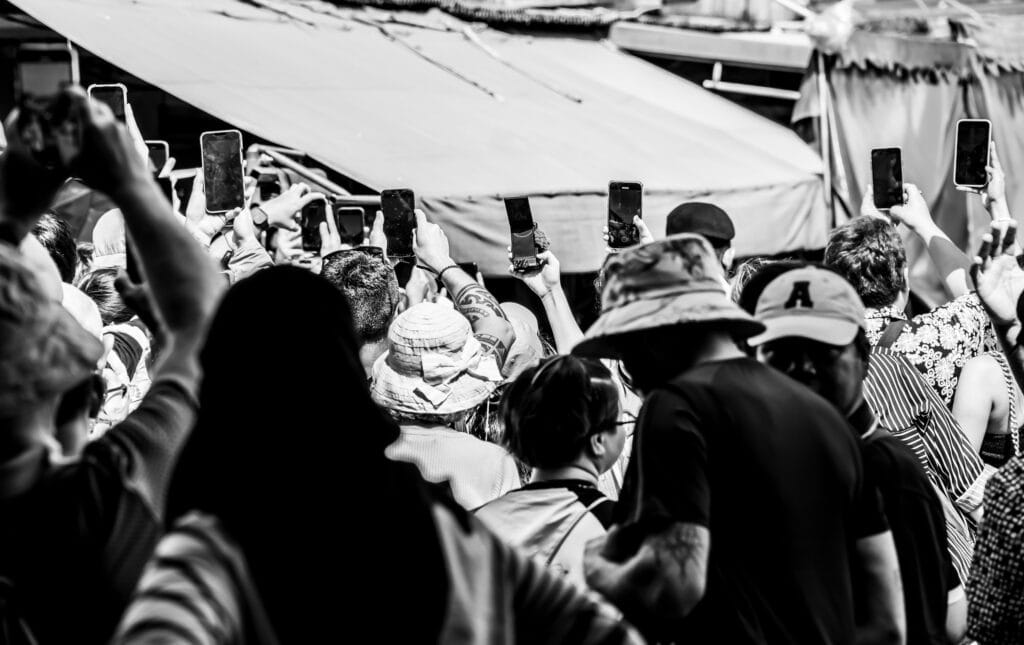 A crowd hold up their mobile phones to photograph the Mae Khlong market train - monochrome