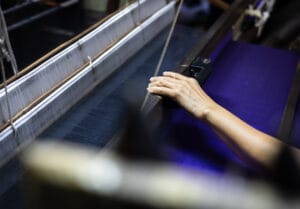 A woman's hand rests on a silk loom mainly loaded with blue thread