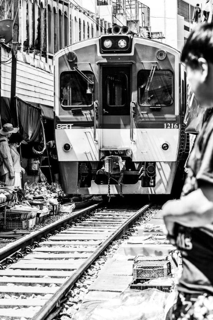 Monochrome low angled view of the Mae Khlong railway market train squeezing alongside people.