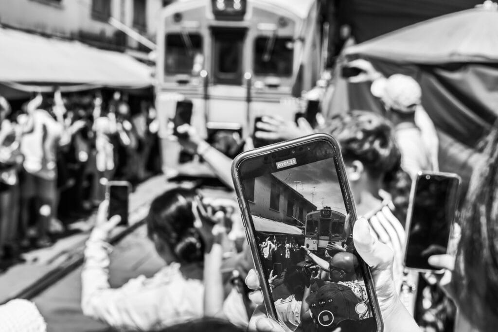 A crowd hold up their mobile phones to photograph the Mae Khlong market train - monochrome