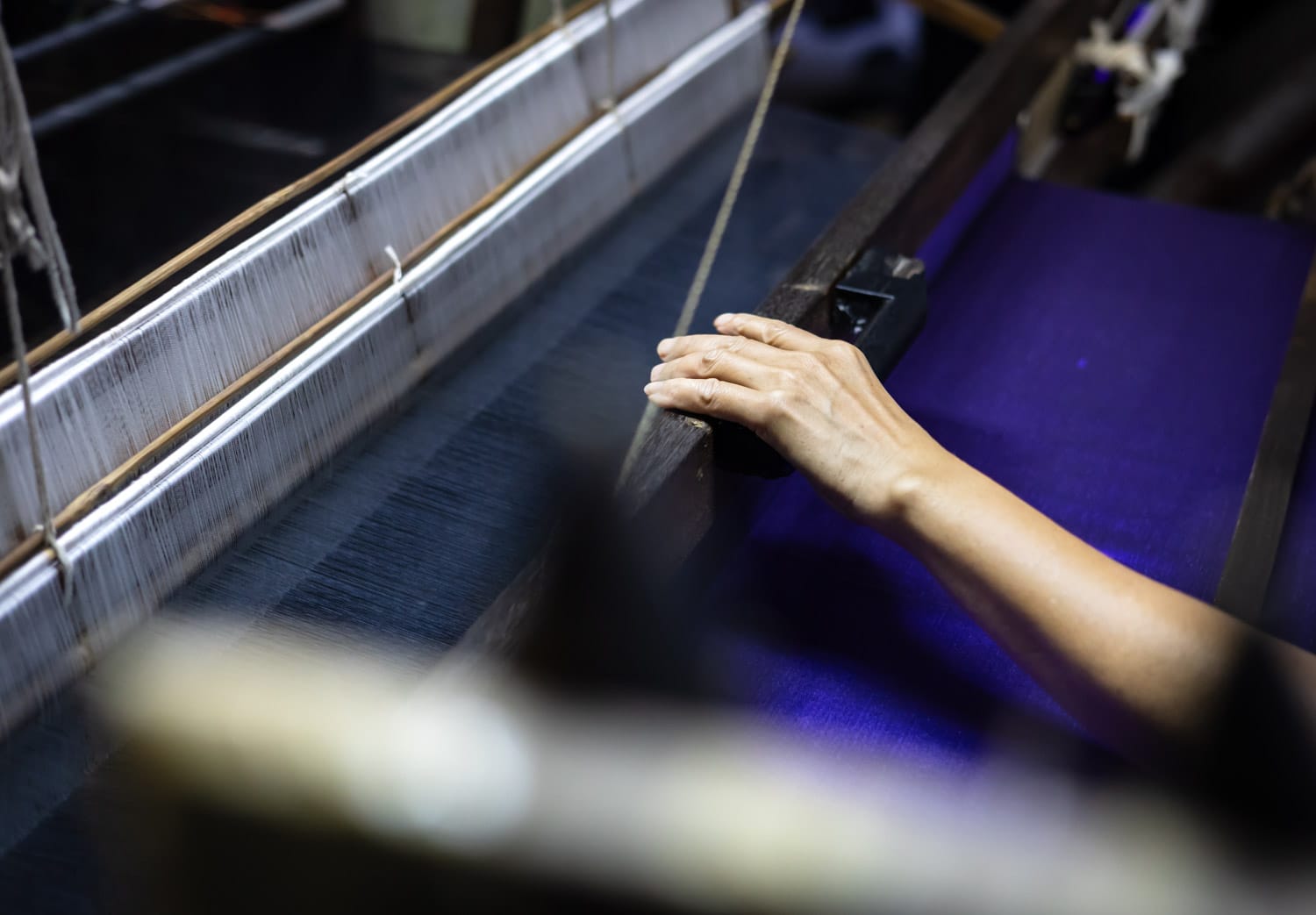 A woman's hand rests on a silk loom mainly loaded with blue thread in Baan Khrua