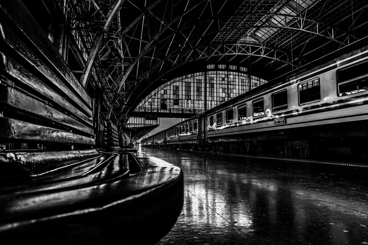 A seat is in the foreground of a view down a platform with a train on the right in Bangkok's Hua Lamphong Railway Station. Monochrome.