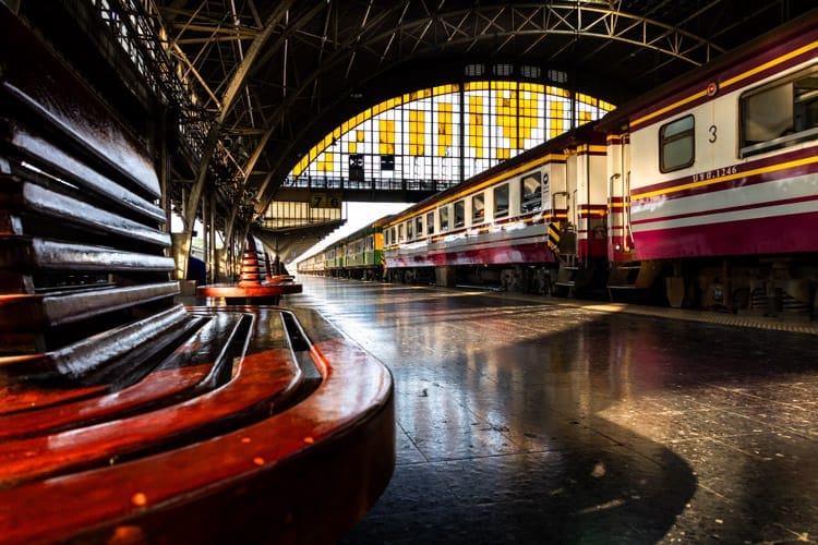The arched stain glass windows of Hua Lamphong Station Bangkok form a backdrop to a train about to depart. In the foreground curved wooden seating catches the late afternoon sun.