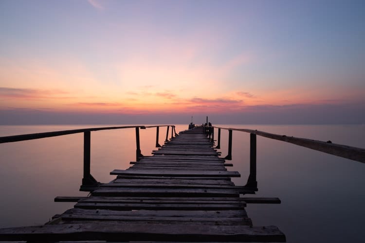 Long exposure photograph of a wooden pier jutting out to sea at sunrise. The sea is silky smooth and the sky is orange and blue. Ao Cho, Koh Samet, Thailand.