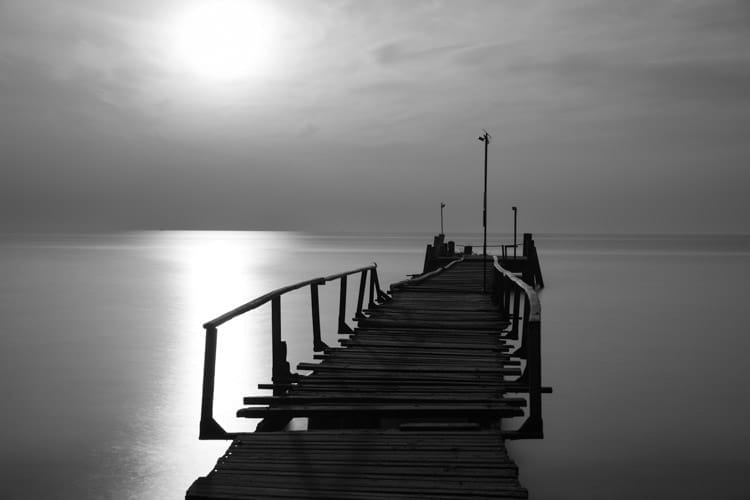 Uneven wooden pier on Koh Samet, extends into the sea. Crooked wooden rails are attached to the sides. Long exposure gives a smooth appearance to the water. Sunlight creates a shimmering path to the horizon. Wispy clouds add to the serene atmosphere.